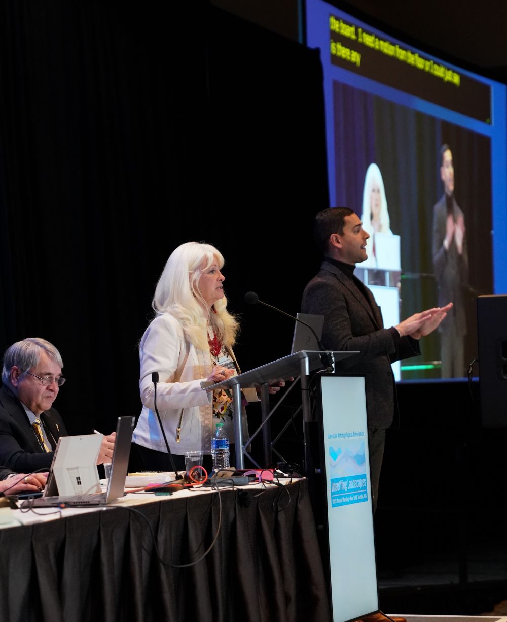 Ramona Pérez stands at a lectern and reads an address from a laptop. She is a light-skinned woman with long white hair wearing a white jacket and a red beaded necklace. To her left is Kyle Duarte, a medium-skinned man with short dark hair. Kyle is wearing a brown suit jacket and a dark turtleneck. Kyle is interpreting Ramona's remarks in American Sign Language. Behind Ramona and Kyle is a projection screen that shows a video feed of the two of them, with captions provided in yellow font at the top of the screen.