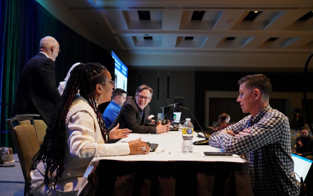 Nate Wambold, a white man with short brown hair, wears a checkered shirt on the right of the frame. He is speaking to AAA president-elect Dr. Whitney Battle-Baptiste, a black woman with braids wearing a white sweater, seated opposite. They are on the stage at the front of a General Session room. Behind them there are several people seated at the head table engaged in conversation.