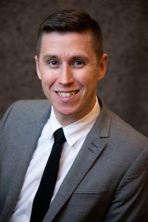 Photo of Nate Wambold, smiling, with a gray blazer, white shirt and black tie, in front of a mottled brown background.
