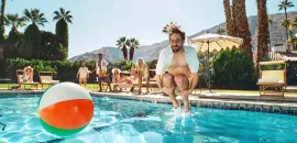 Photo of man jumping into a pool with a beach ball nearby.