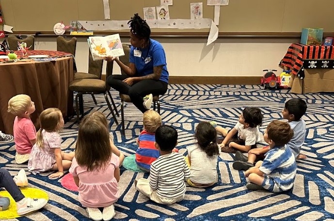 Adult in a blue shirt sitting in a chair and instructing 10 children sitting on a carpet in front of her.