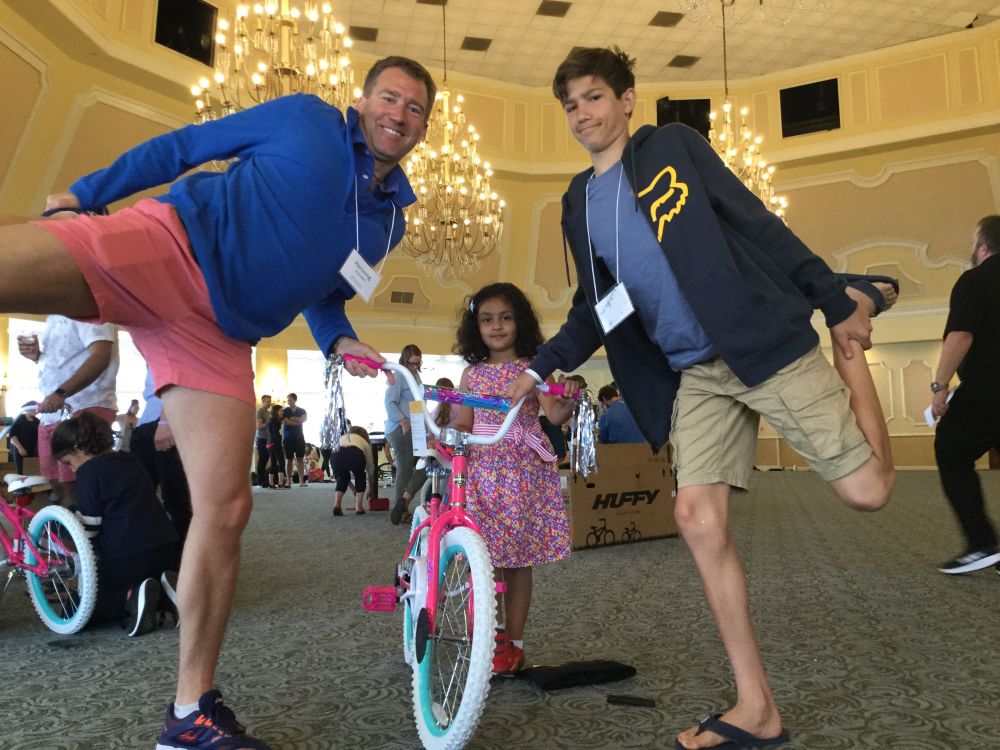 Photo of two adults with their legs lifted to the side, with a girl in the middle with a bike.