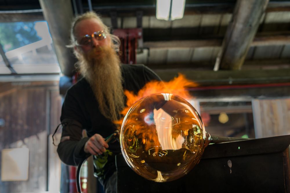 Photo of a bearded man blowing a glowing ball of glass.