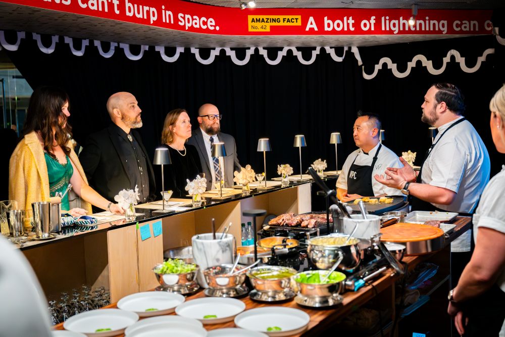 Photo of a group of chefs preparing food in front of four diners.