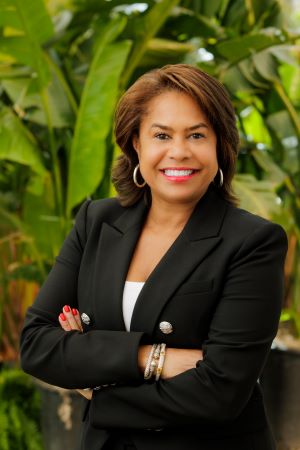 Photo of Julie Coker, standing with her arms crossed, in front of green foliage.