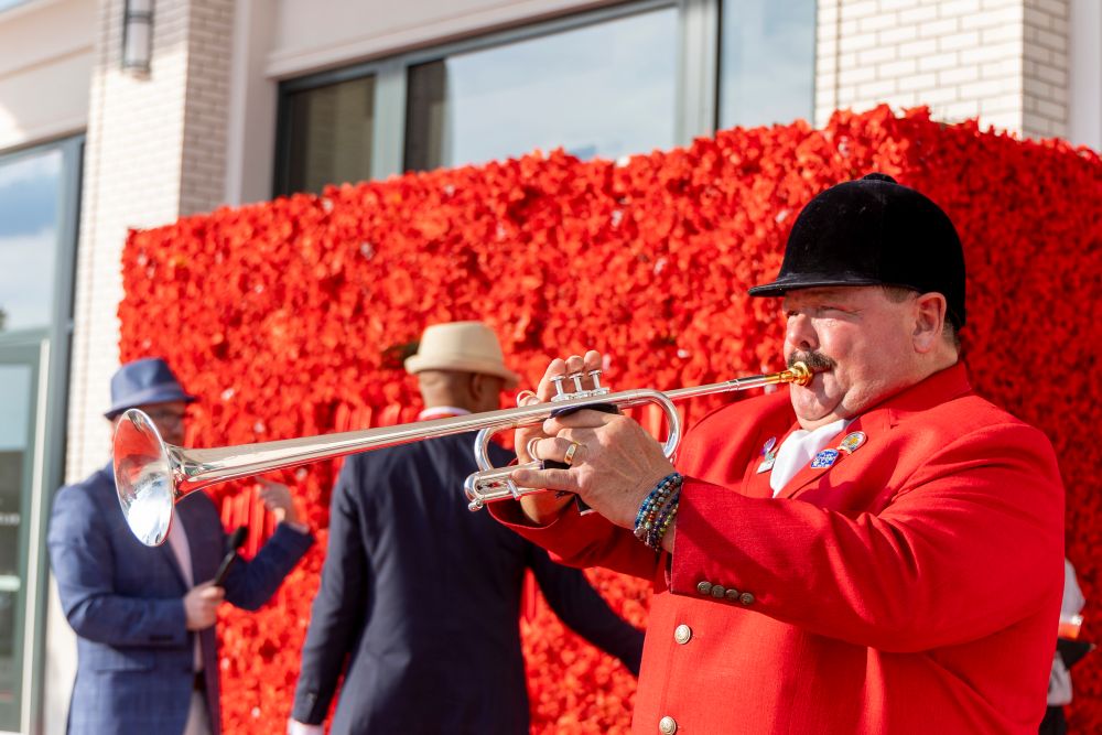 Photo of man in red jacket and black hat blowing a long trumpet in front of a hedge of red flowers.