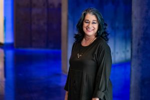 Photo of Maura Gast in a black dress in front of a blue background.