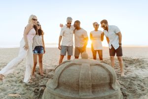 Six people standing around a sandcastle on a beach.