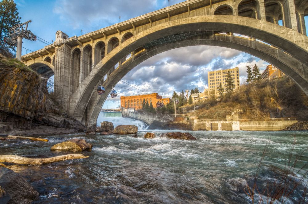 Photo of downtown Spokane from the Spokane River, with a large arched bridge in the background.