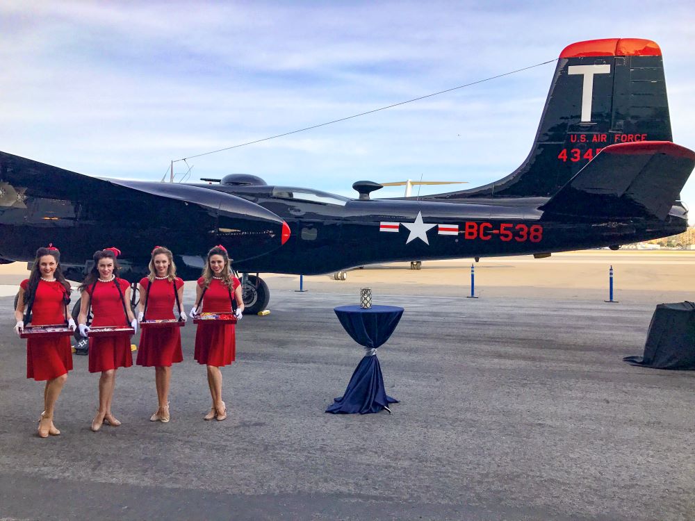 Photo of a vintage World War II-era plane with four stewardesses dressed in vintage red dresses in front, holding trays.