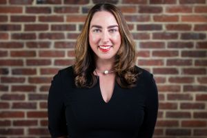 Photo of Tara Miller, in a black shirt in front of a red brick wall.