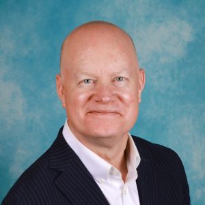 Photo of Tom Noonan in a black suit and white shirt in front of a blue background.