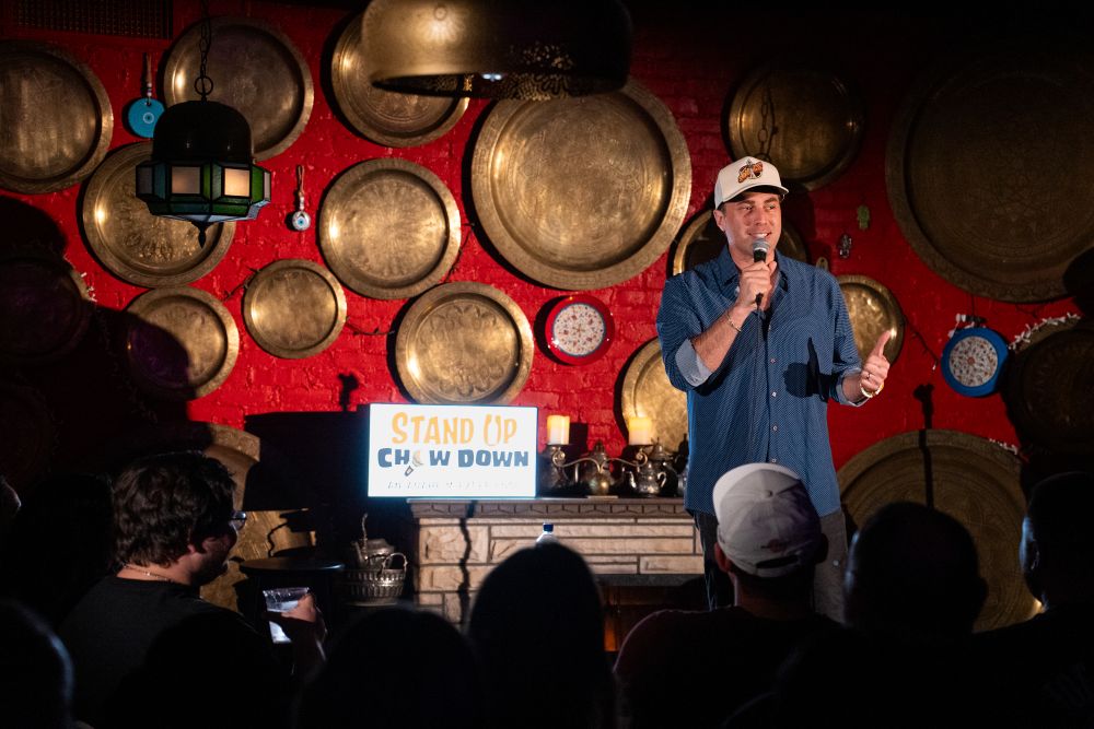 Photo of comedian on stage, with copper Moroccan plates in the background.