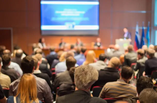 Photo of meeting room full of attendees, listening to a presenter.