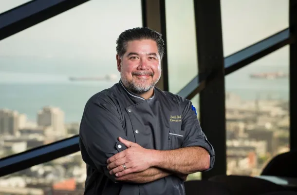 Photo of Chef David Hollands standing in front of a picture window with the skyline of San Francisco in the background.