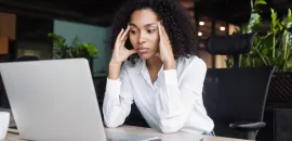 A business woman in a white blouse clasping her heads to her head in frustration, over a laptop.