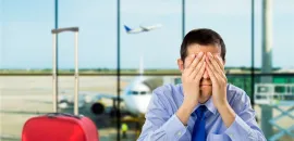 Photo of a man in an airport with his hands on his face, exasperated, with a red piece of carry on luggage next to him.