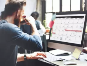 Man drinking water working on a computer with a calendar displayed on it.