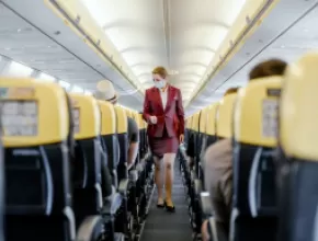 Masked flight attendant walking down airline aisle.