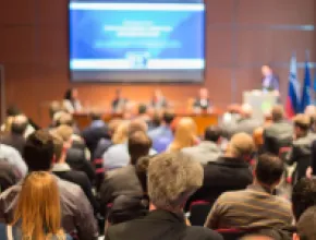 Photo of meeting room full of attendees, listening to a presenter.