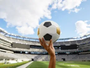 Image of a hand holding a soccer ball with a stadium in the background.