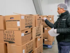 Image of a man standing in front of boxes of donated food from Chefs to End Hunger