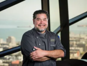 Photo of Chef David Hollands standing in front of a picture window with the skyline of San Francisco in the background.