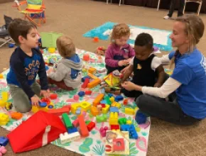 Image of an adult sitting on a carpet with four children and toys.