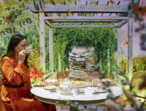 Photo of a woman in an orange dress sipping tea at a table in a garden setting.