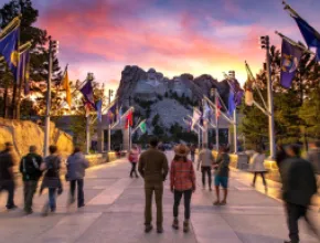 People standing in a walkway looking at Mt. Rushmore, with flags on the sides.
