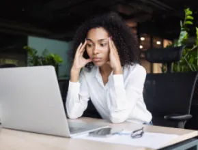 A business woman in a white blouse clasping her heads to her head in frustration, over a laptop.