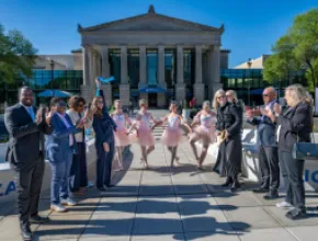 A crowd of people stand in front of a historic building, with ballet dancers, to celebrate cutting the ribbon.