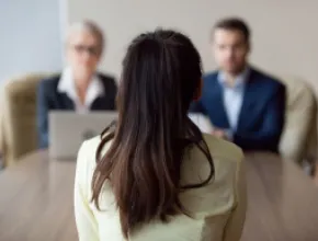 Image of two people interviewing a job candidate at a desk, with the candidate's back to the viewer.