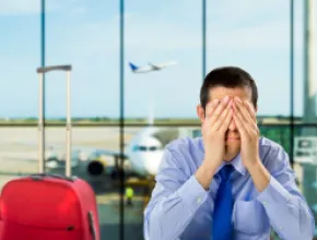 Photo of a man in an airport with his hands on his face, exasperated, with a red piece of carry on luggage next to him.