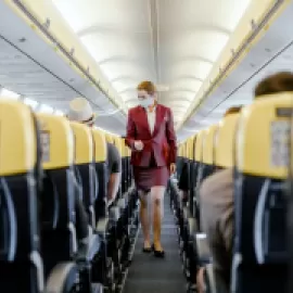 Masked flight attendant walking down airline aisle.