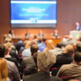 Photo of meeting room full of attendees, listening to a presenter.