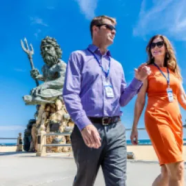 Man and woman attendees walking with King Neptune statue in the background in Virginia Beach