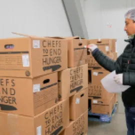 Image of a man standing in front of boxes of donated food from Chefs to End Hunger