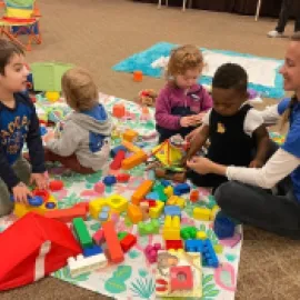 Image of an adult sitting on a carpet with four children and toys.
