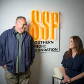Photo of Chris Shepherd (left, standing) and Lindsay Brown (right sitting) in front of a Southern Smoke Foundation logo.