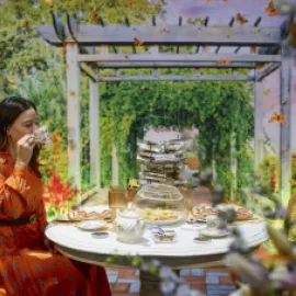 Photo of a woman in an orange dress sipping tea at a table in a garden setting.