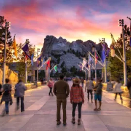 People standing in a walkway looking at Mt. Rushmore, with flags on the sides.