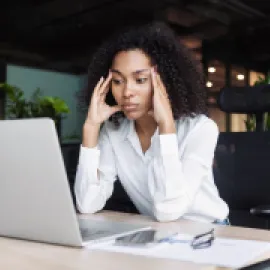 A business woman in a white blouse clasping her heads to her head in frustration, over a laptop.