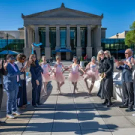 A crowd of people stand in front of a historic building, with ballet dancers, to celebrate cutting the ribbon.