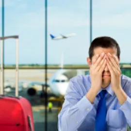 Photo of a man in an airport with his hands on his face, exasperated, with a red piece of carry on luggage next to him.