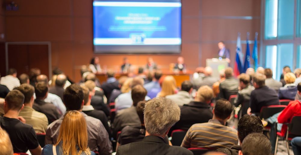 Photo of meeting room full of attendees, listening to a presenter.