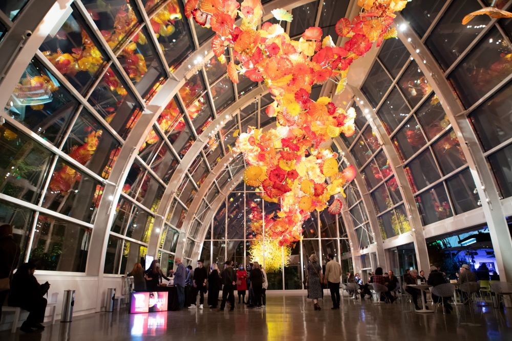 Photo of people gathered in a hall for Refract, under a glowing glass sculpture.