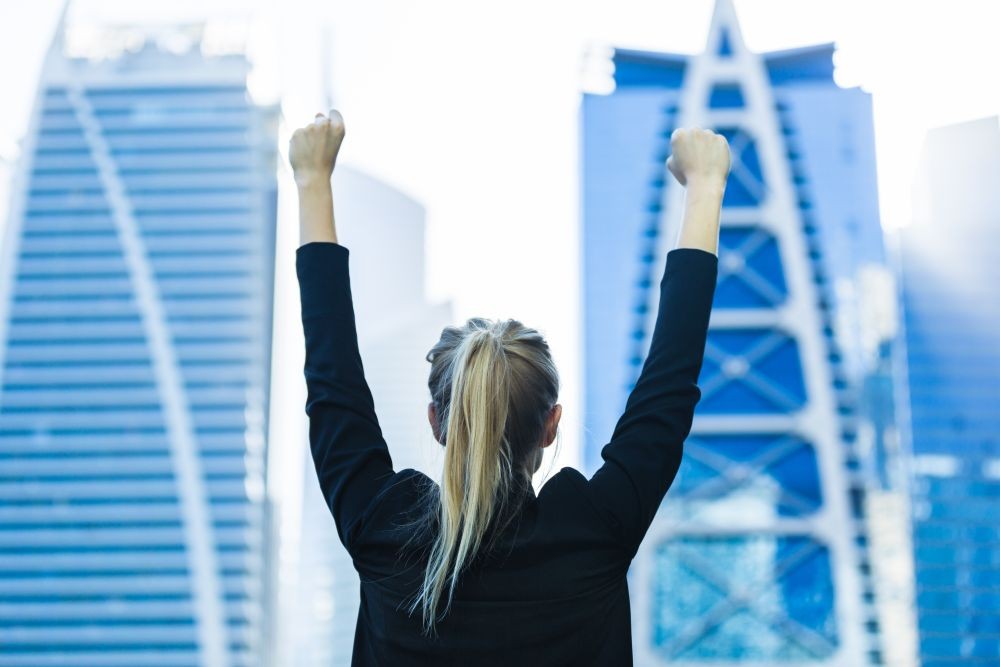 Photo of a woman with her arms up in the air in front of two skyscrapers.