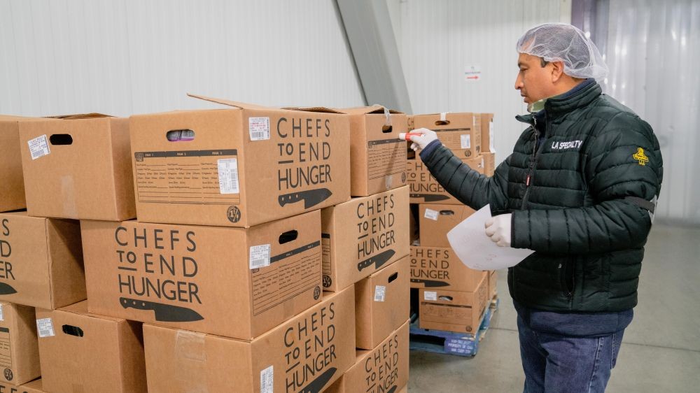 Image of a man standing in front of boxes of donated food from Chefs to End Hunger