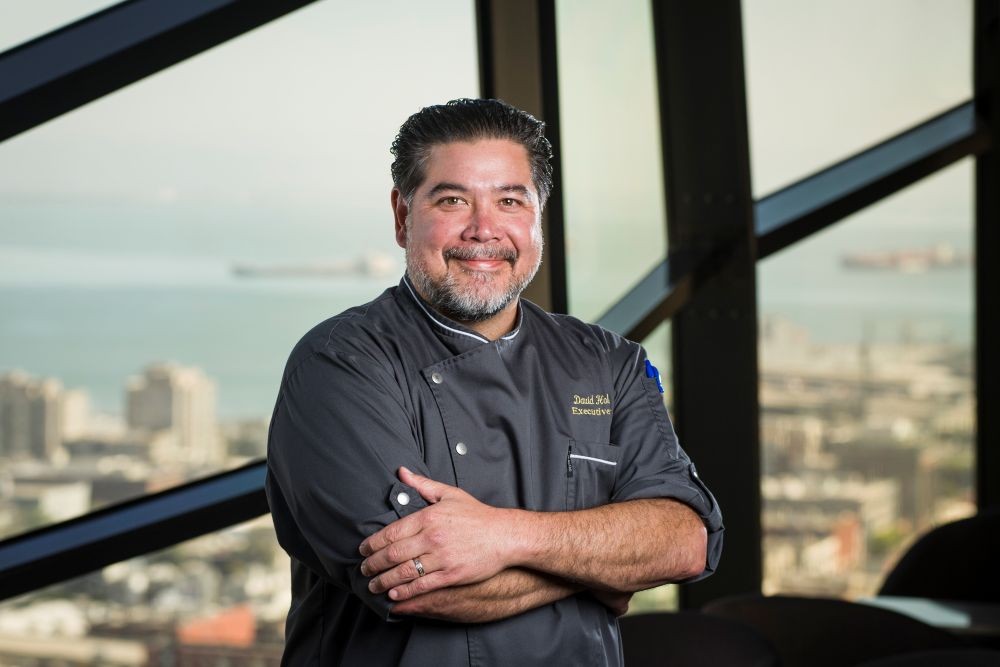 Photo of Chef David Hollands standing in front of a picture window with the skyline of San Francisco in the background.